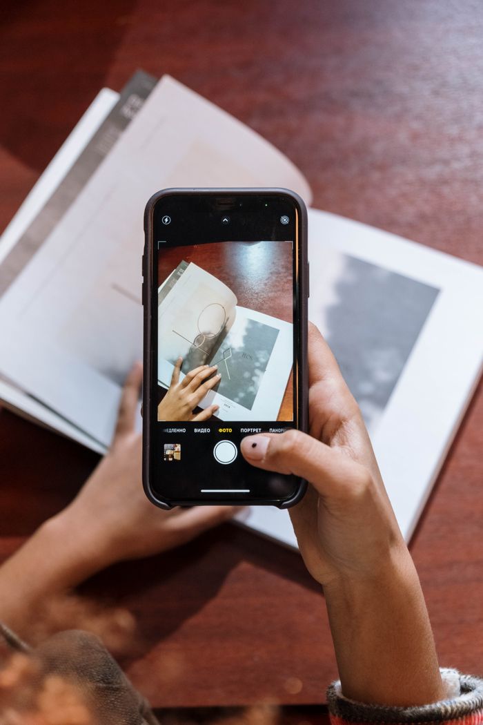 Services Person photographing a book with a smartphone indoors on a wooden table.
